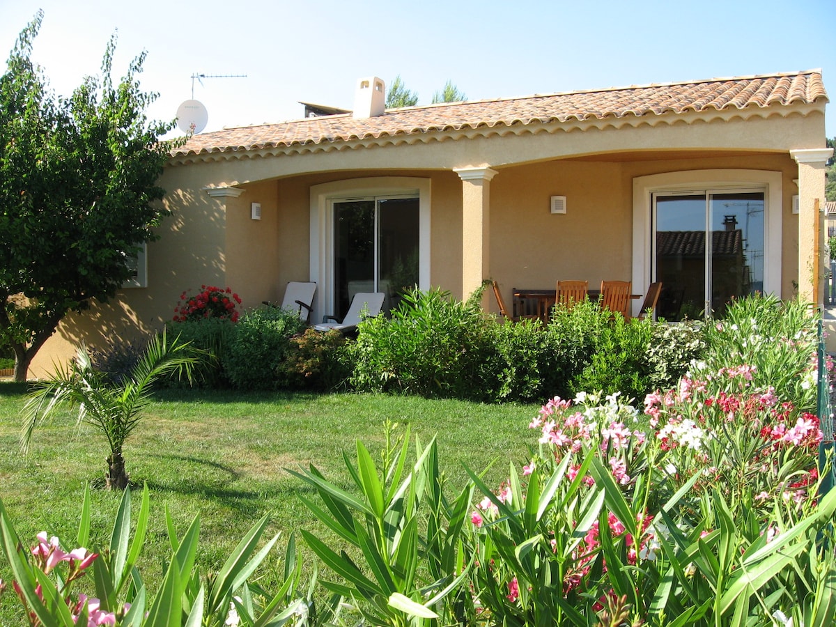 The villa is framed by vibrant greenery and blooming flowers, showcasing a sunlit facade. Two lounge chairs sit on a small terrace, accompanied by a wooden dining set. The expansive lawn is punctuated by well-maintained shrubs, all under a clear blue sky.