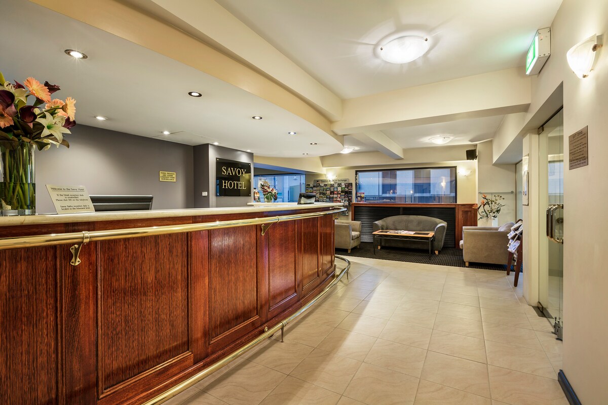 The hotel reception area features a polished wooden front desk, complemented by subtle lighting. A cozy seating area with soft couches and a vase of flowers adds warmth. Shelving in the background displays various items, enhancing the space's welcoming ambiance.
