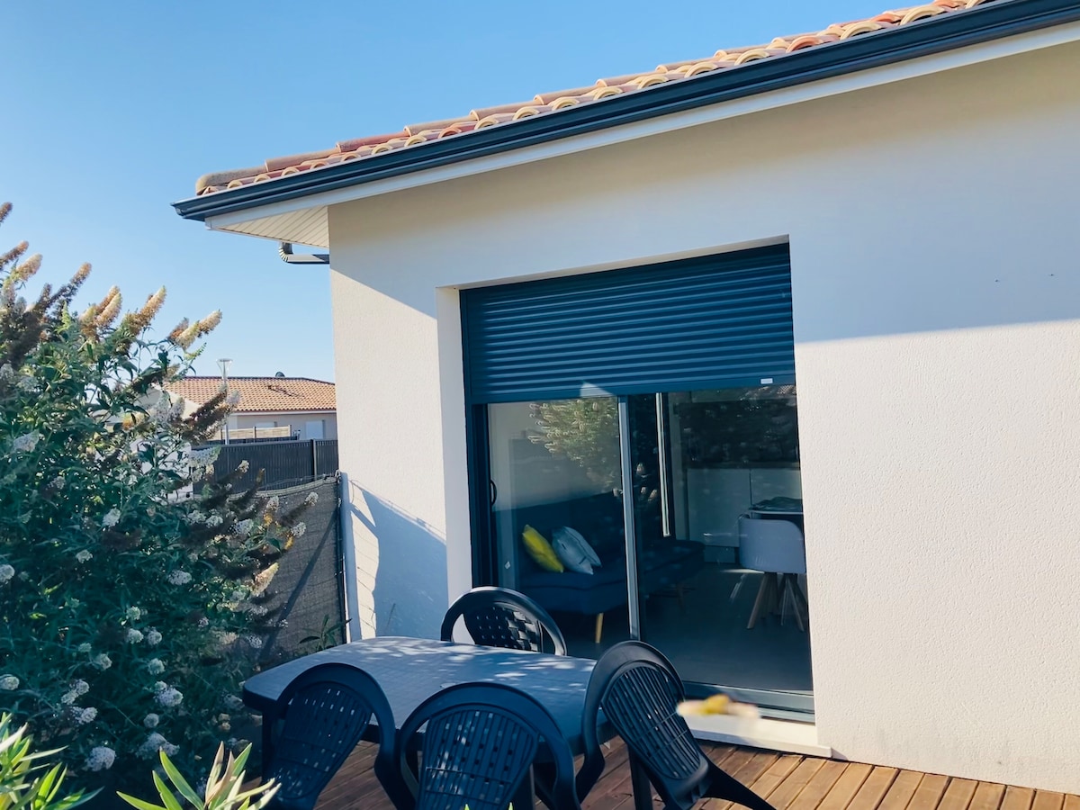 An outdoor terrace showcases a dining table surrounded by black chairs, bordered by green shrubs. Large doors lead into a bright interior, where a seating area with cushions is visible. The space benefits from natural light and overlooks the picturesque surroundings.