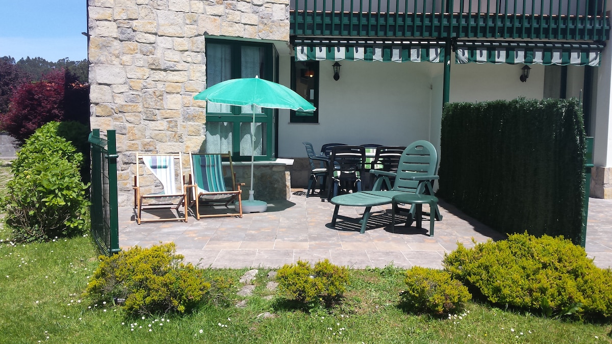 An outdoor patio is presented with a stone flooring surface. Comfortable lounge chairs and a sun umbrella are arranged around a dining table. A green hedge provides privacy, and well-maintained shrubs are visible, enhancing the inviting outdoor space.