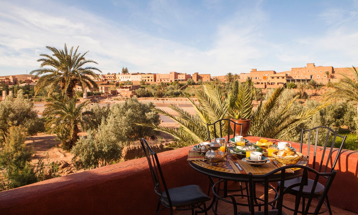 A dining table set for breakfast is placed on a balcony, offering views of palm trees and traditional buildings. Plates and cups are arranged neatly, with a picturesque natural landscape in the background. The warm sunlight illuminates the scene, creating a serene outdoor setting.