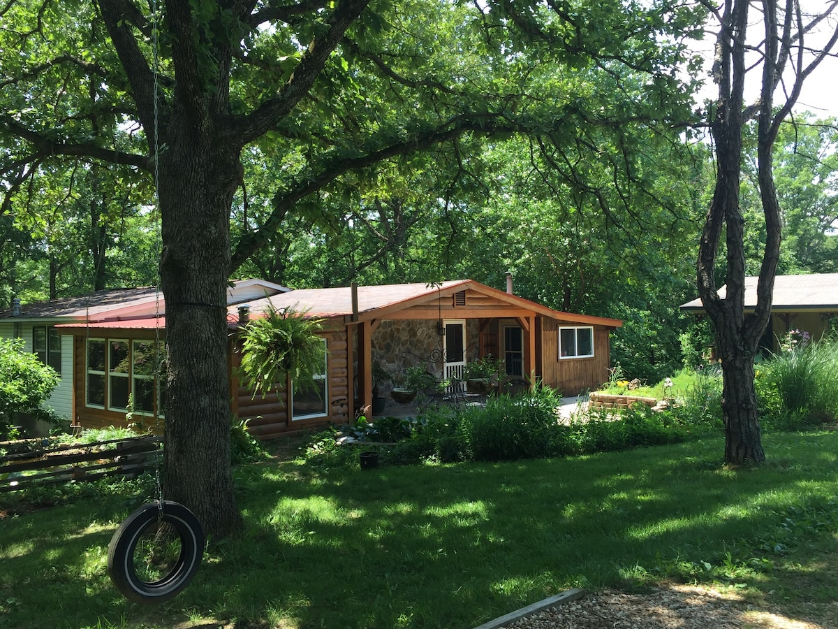 A secluded cottage is surrounded by lush greenery and tall trees. The inviting exterior features a combination of wood and stone elements, with a porch visible at the entrance. A tire swing hangs from a nearby tree, adding to the tranquil ambiance of the yard.