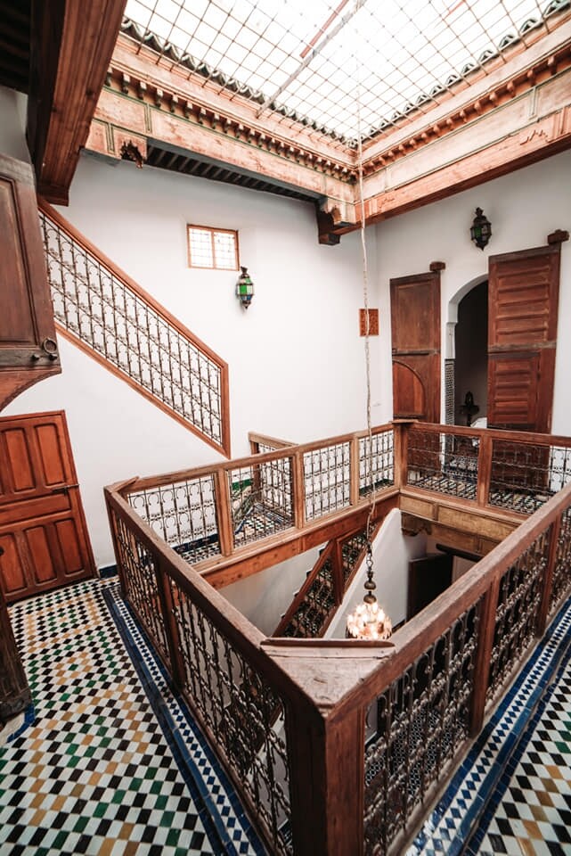 An elegant interior features a central atrium with a glass skylight, flooding the space with natural light. Ornate wooden railings surround the staircase, complemented by intricately patterned ceramic tiles on the floor. Wooden doors lead to additional spaces, enhancing the traditional aesthetic.