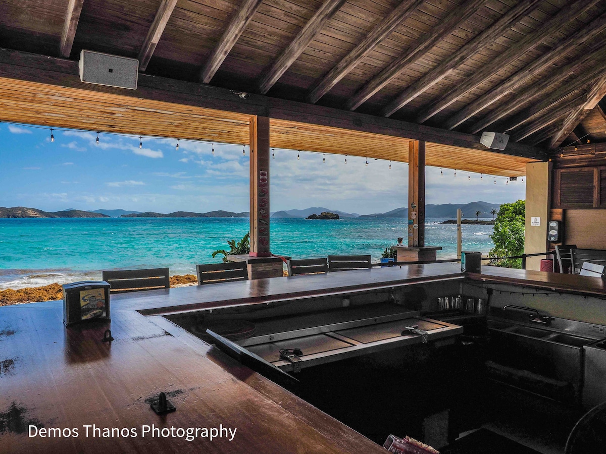 A wooden bar area features a rustic countertop, overlooking a breathtaking view of the turquoise waters and distant islands. The scene is illuminated by soft natural light, with comfortable seating visible along the bar. The open space invites relaxation and enjoyment of the beachfront atmosphere.