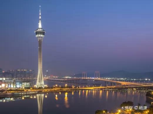 The image captures the Macau Tower illuminated at night, with its distinctive height prominently framed against a twilight sky. The surrounding water reflects the tower's lights, while the bridge nearby adds to the scenic cityscape.
