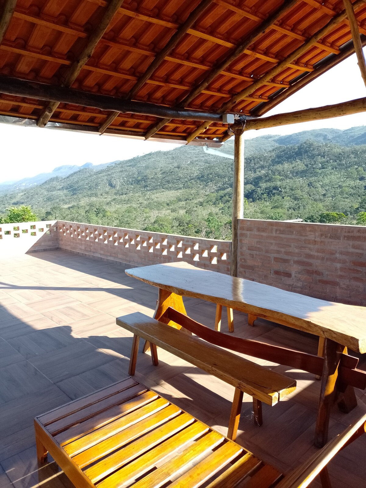 An outdoor terrace features a wooden table with matching chairs, positioned to take in sweeping views of the surrounding mountains. A clay brick wall provides a rustic feel, while the sloped roof adds character to the open space, inviting relaxation in nature.