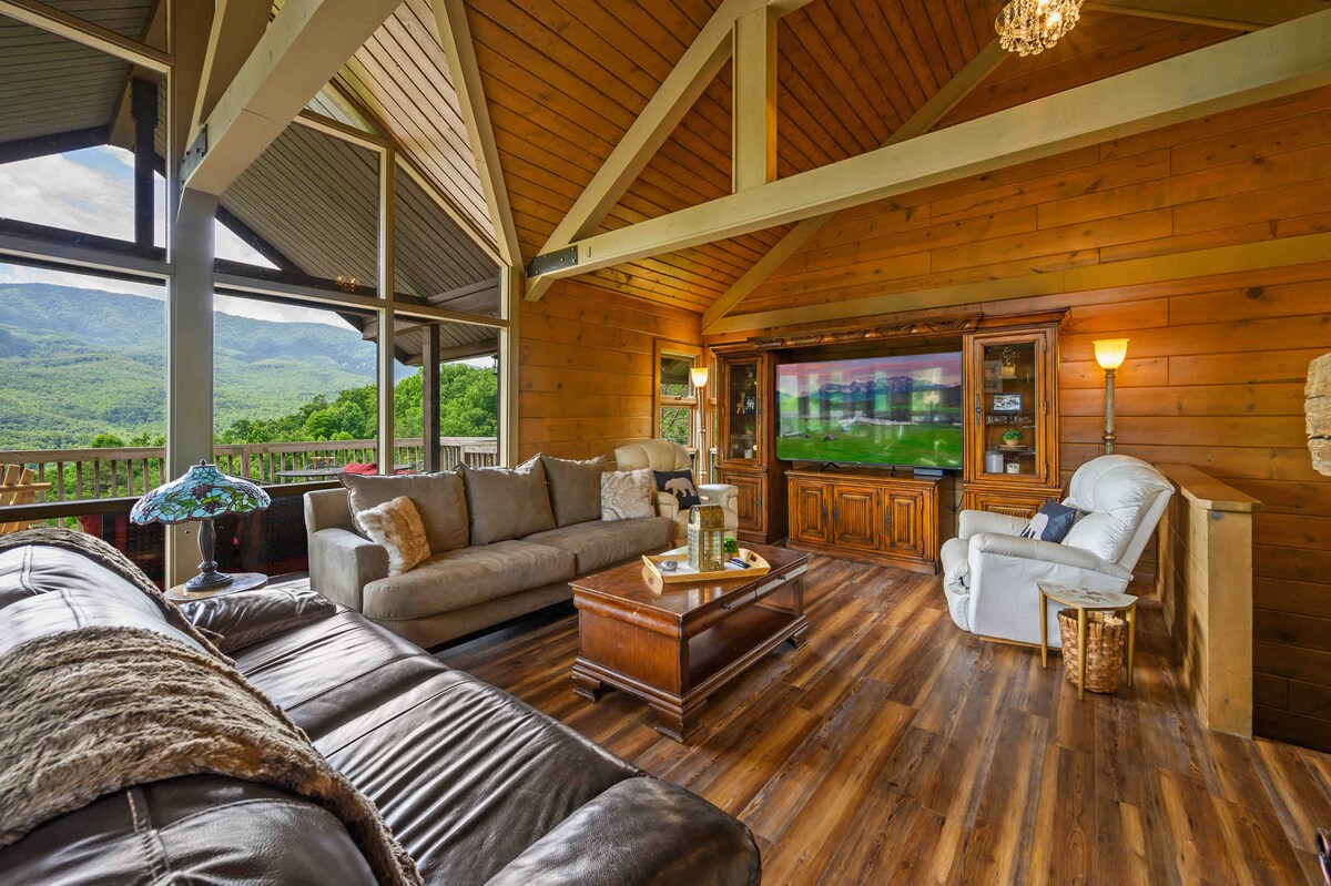 The living room features two spacious couches and a recliner, arranged around a wooden coffee table. A large TV is mounted on the wall, complemented by a wooden cabinet. Expansive windows offer views of the surrounding mountains, while natural light fills the space.
