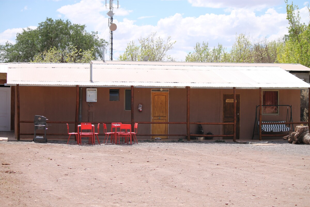 A single-story building is shown with a metal roof and earth-toned exterior. Red chairs and a table are arranged outside, alongside a grill. Surrounding greenery and an open area enhance the setting, with additional wooden structures visible in the background.