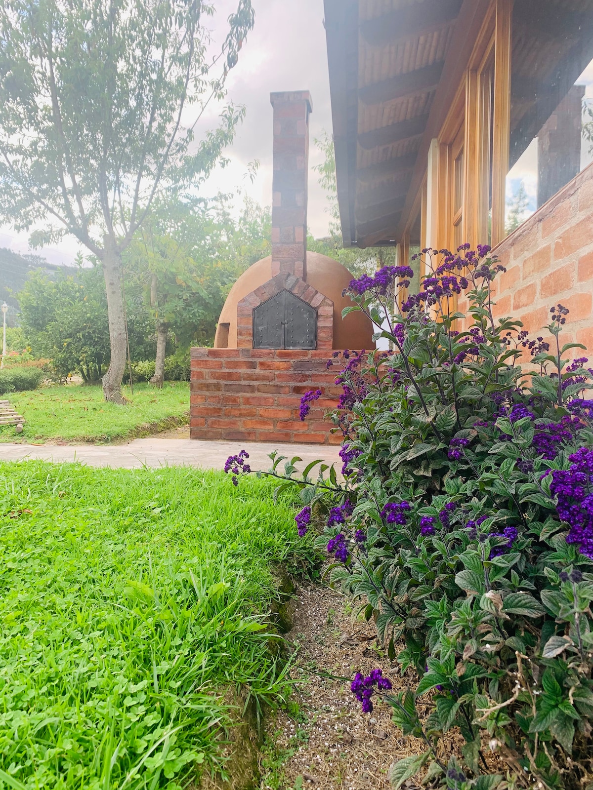 A traditional brick oven is seen beside a well-maintained garden, where vibrant purple flowers bloom among lush green grass. Trees provide a natural backdrop, enhancing the serene outdoor setting.