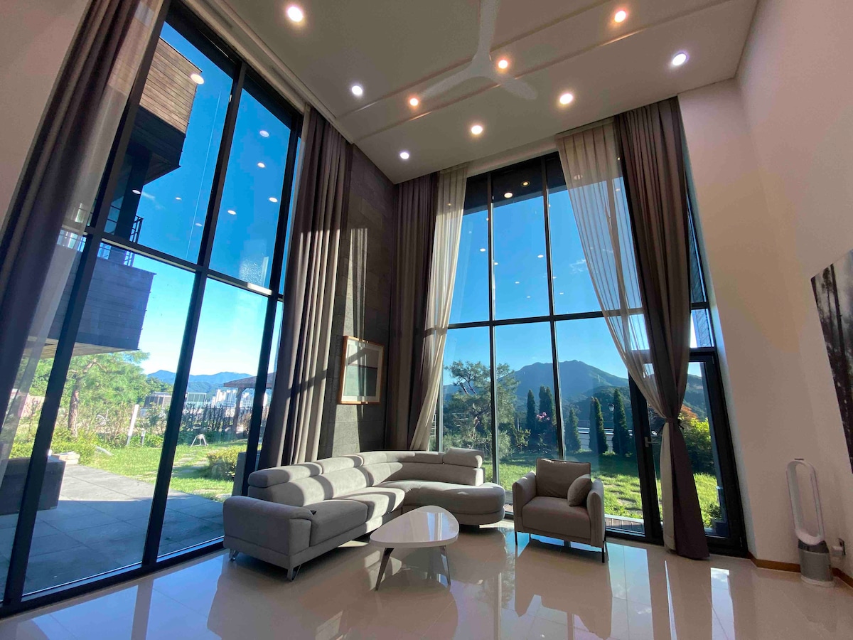 A modern living area features tall windows that offer views of the surrounding landscape and mountains. Comfortable seating is arranged around a sleek coffee table, with soft lighting enhancing the spacious interior. The neutral color palette complements the natural light entering through the glass.