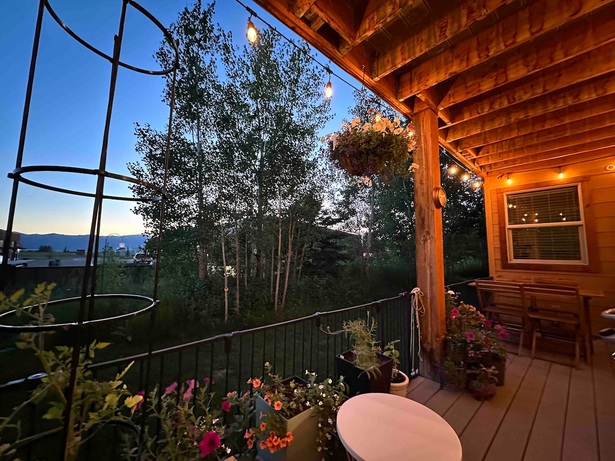 The outdoor deck is adorned with plants and flowers, complemented by string lights creating a soft glow. A small dining table and chairs offer a cozy seating area. Quaking aspens frame the view, providing a peaceful backdrop as evening falls.