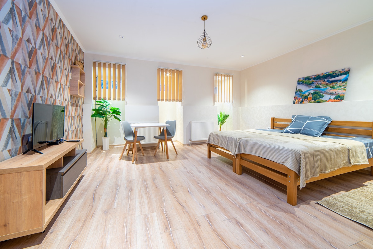 A spacious room features a comfortable bed with a simple beige blanket and decorative pillows. A small dining area with two chairs and a table is positioned nearby. Natural light filters through angled blinds on two windows, complemented by green plants and a television on a wooden stand.