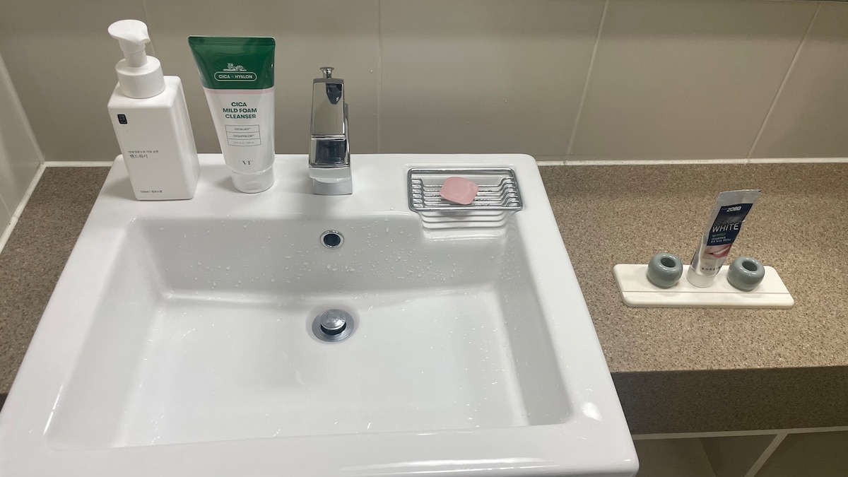 A clean bathroom sink area is displayed, featuring a white porcelain sink with modern fixtures. Various toiletries are arranged, including a soap dispenser, a cleansing foam tube, and a small soap dish. The countertop is a light brown material, complemented by a neatly organized soap holder.