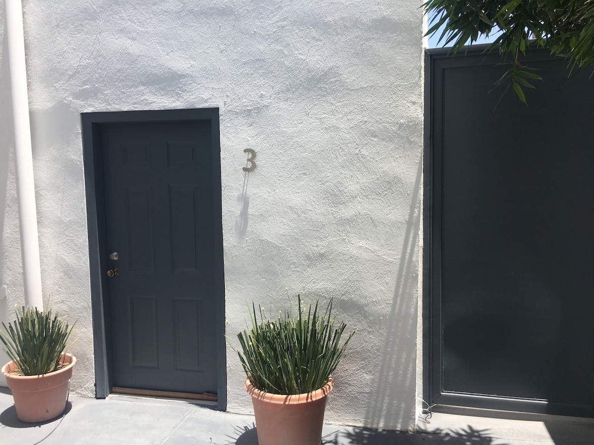 A dark blue door labeled '3' is set within a light-colored wall. On either side of the door, potted plants with slender green leaves are positioned, enhancing the entryway. A blank wall section on the right showcases a smooth surface, contributing to the minimalistic aesthetic.