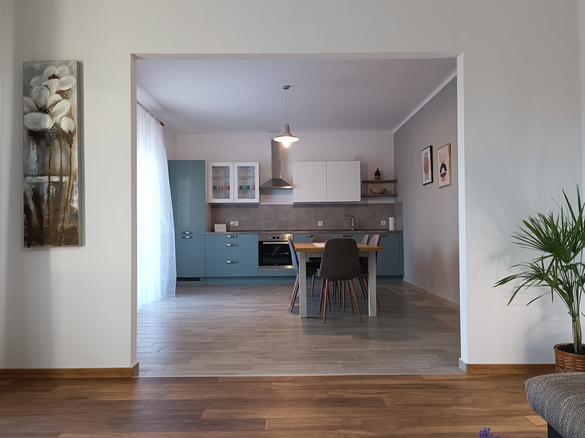 A modern kitchen and dining area are visible through an archway, featuring teal cabinetry and a wooden dining table surrounded by chairs. Natural light enters through a window, while decorative artwork adorns the walls, creating a bright and inviting space.