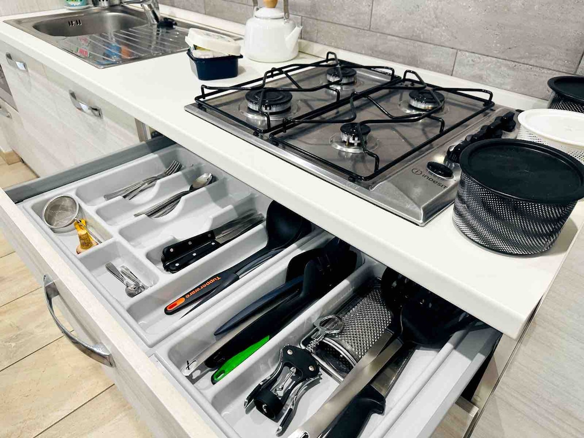 A modern kitchen is depicted, featuring a gas stove with four burners above a pristine countertop. Below, a neatly organized drawer reveals a variety of cooking utensils, including knives, graters, and measuring tools, all arranged for easy access.