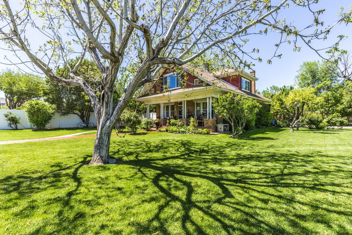 A two-story home is surrounded by a large grassy yard, with several trees providing shade. Sunlight filters through the leaves, casting intricate shadows on the lawn. The front porch invites relaxation, while the surrounding landscape features an array of greenery.