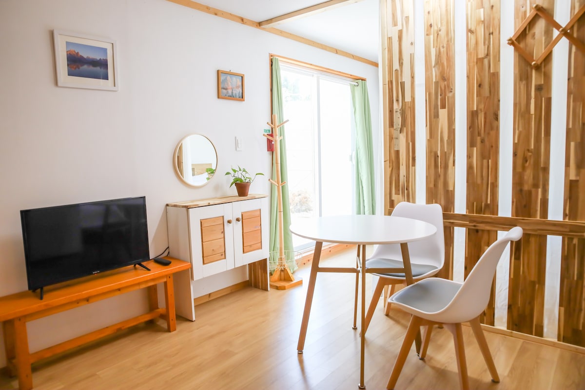 A bright living space features a round dining table accompanied by two chairs. A small wooden cabinet and a flat-screen TV are positioned against the wall. Large windows allow natural light to fill the room, which has a warm wooden floor and decorative wooden paneling.