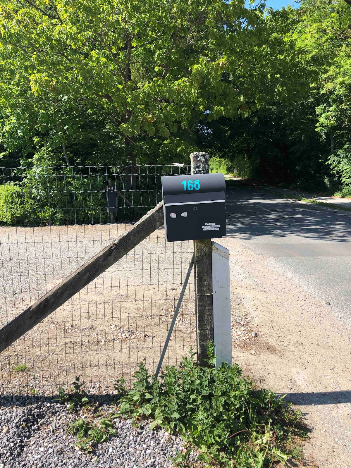 A modern black mailbox with the number '168' is positioned on a wooden post at the entrance of a gravel pathway. Lush greenery surrounds the mailbox, with a fence visible in the background, and a quiet road leading away from the property.