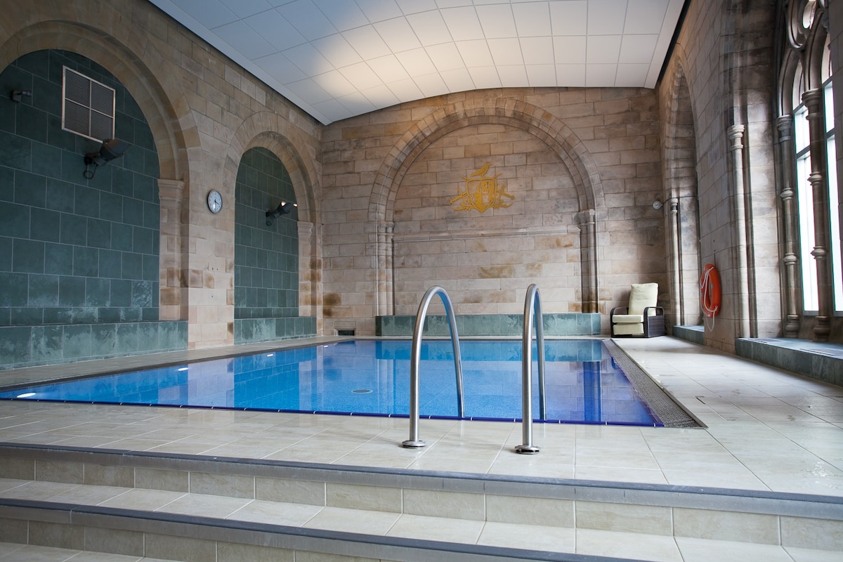 An indoor heated swimming pool is framed by stone walls featuring arched windows, allowing natural light to illuminate the space. The pool is equipped with steps for easy access, and a relaxation area is visible to one side.