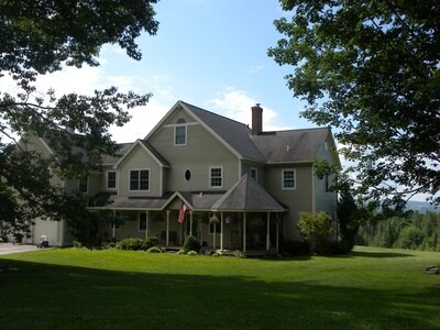 The exterior of the inn is displayed, showcasing a multi-story structure with a covered porch. Surrounding greenery and trees create a serene environment, while a flag is visible near the entrance. The backdrop features a clear sky and distant hills.