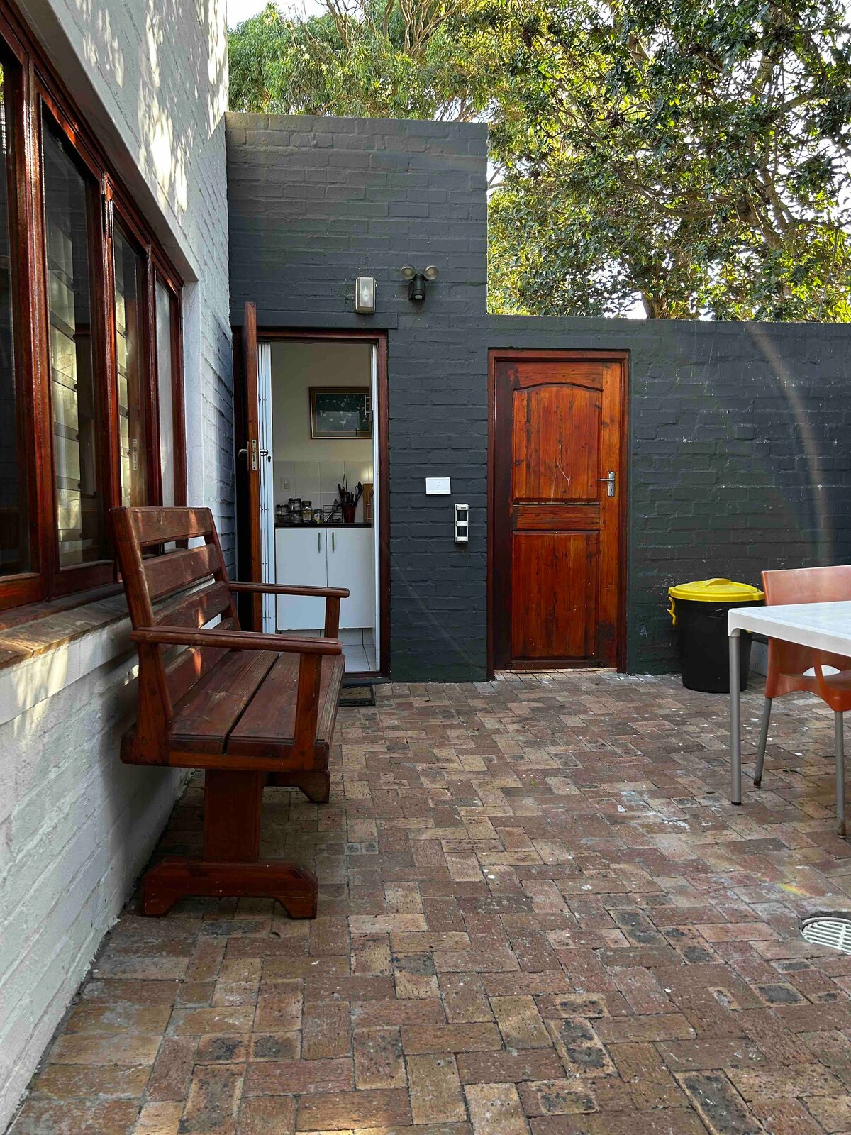 A small courtyard is featured, showcasing rustic brick flooring and a wooden bench. A door leads to a kitchen area, while a wooden door and a contrasting trash bin are visible on one side. Natural light filters through the surrounding greenery.