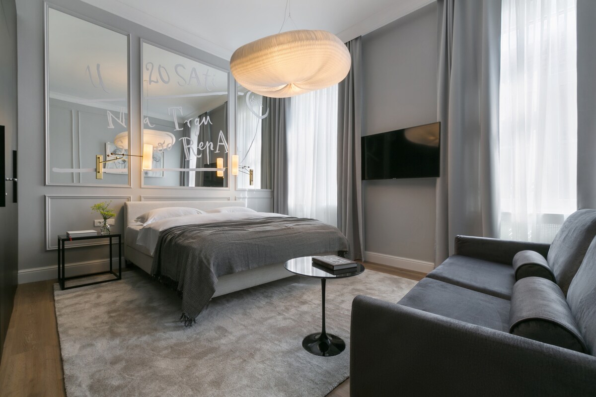 A spacious studio apartment features a king-sized bed with a gray blanket and decorative pillows. Natural light filters through sheer curtains beside a large mirror. A comfortable sofa sits near a round coffee table, while a flat-screen TV is mounted on the wall.