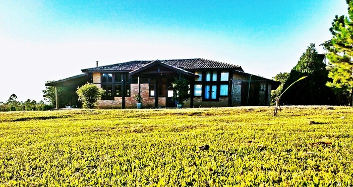 A spacious wooden house is surrounded by lush green grass, showcasing large windows that frame the natural light. The architecture reflects a blend of rustic charm and modern design, inviting guests to enjoy the serene landscape of the countryside.