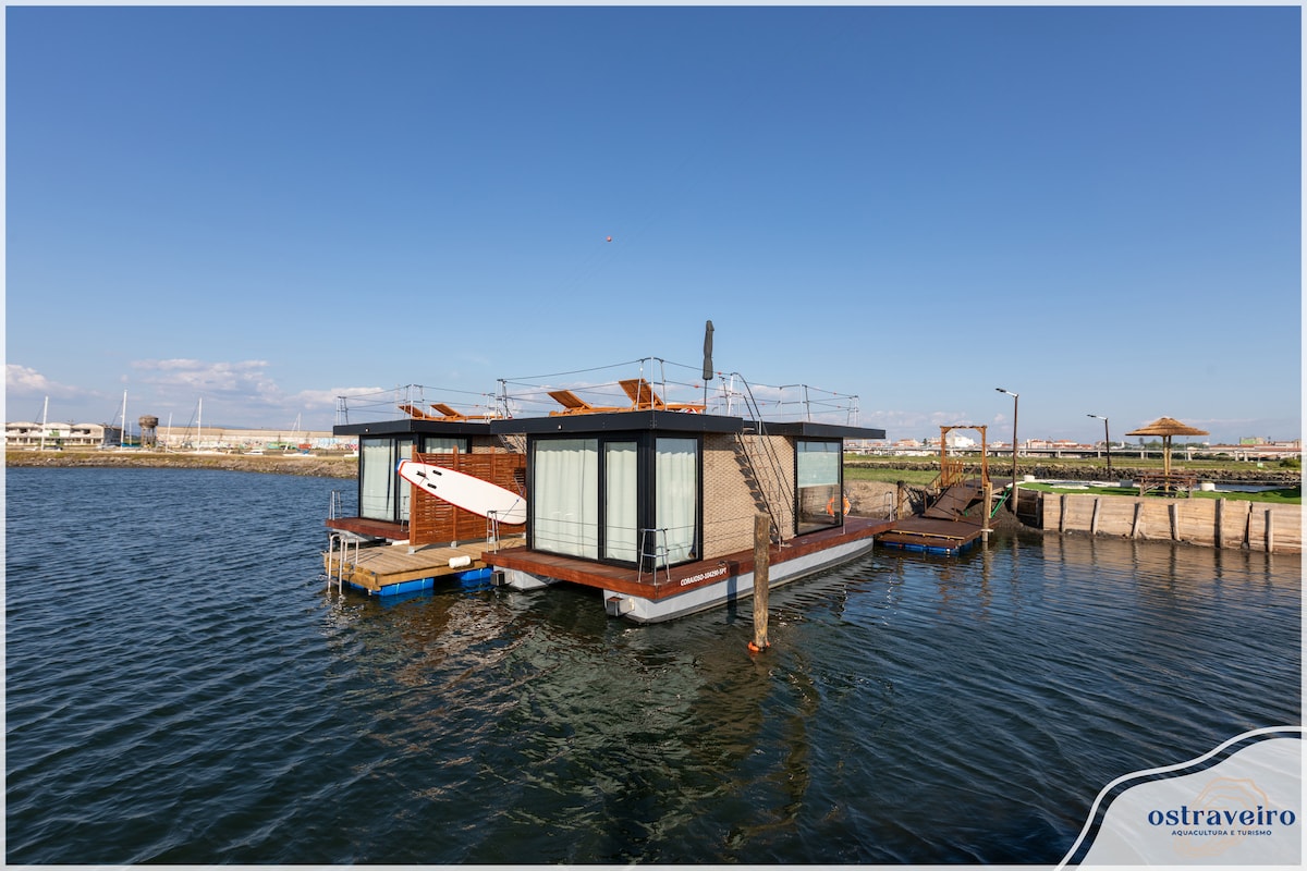 A modern floating house is anchored in calm waters, featuring glass walls that provide views of the surroundings. A wooden deck extends from the structure, with outdoor seating and a surfboard resting against the wall. The clear blue sky complements the tranquil setting.