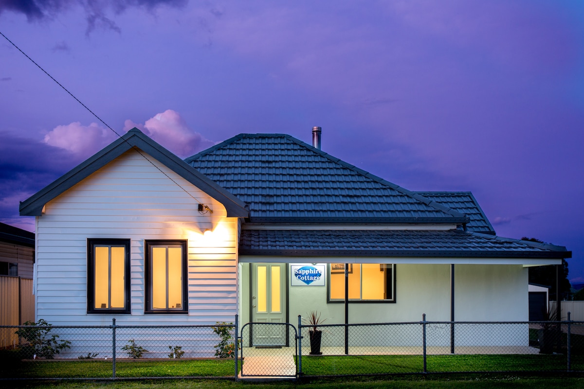 The exterior of Sapphire Cottage is presented, showcasing a single-story structure with a pitched roof. Soft evening light illuminates the entrance, while two windows are positioned symmetrically. A fenced front yard features a well-maintained lawn, enhancing the inviting appearance of the home.