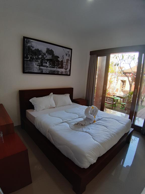 A bright bedroom features a spacious bed with a white comforter and decorative towel art shaped like a swan. Natural light filters through large glass doors that open to a view of outdoor seating and greenery. A framed black-and-white photograph adorns the wall over the bed.