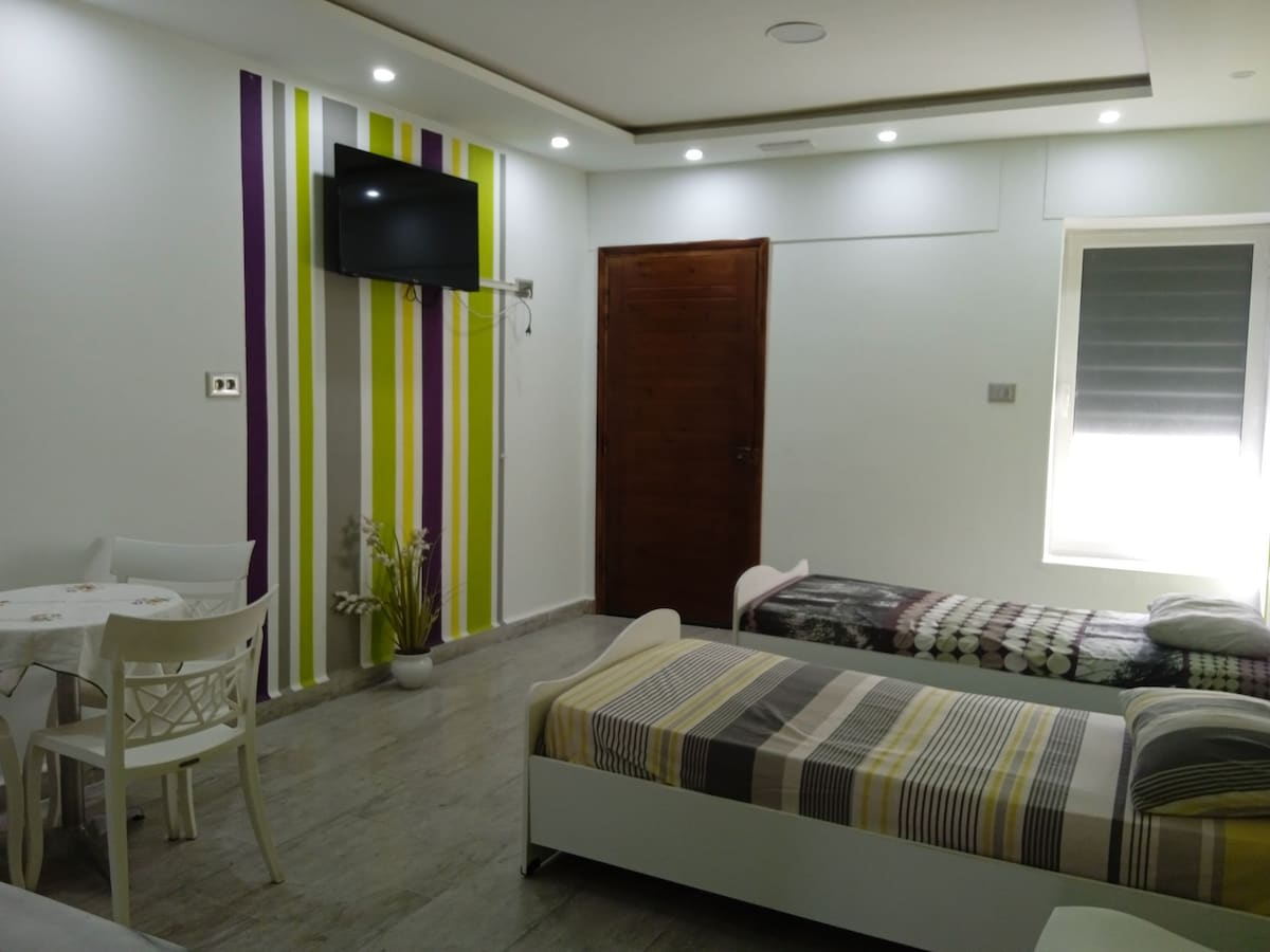 A modern guest room features two beds with striped bedding, complemented by a small table and chairs. Colorful vertical stripes adorn one wall, and a wall-mounted television is visible. Natural light filters through a window with closed shades.