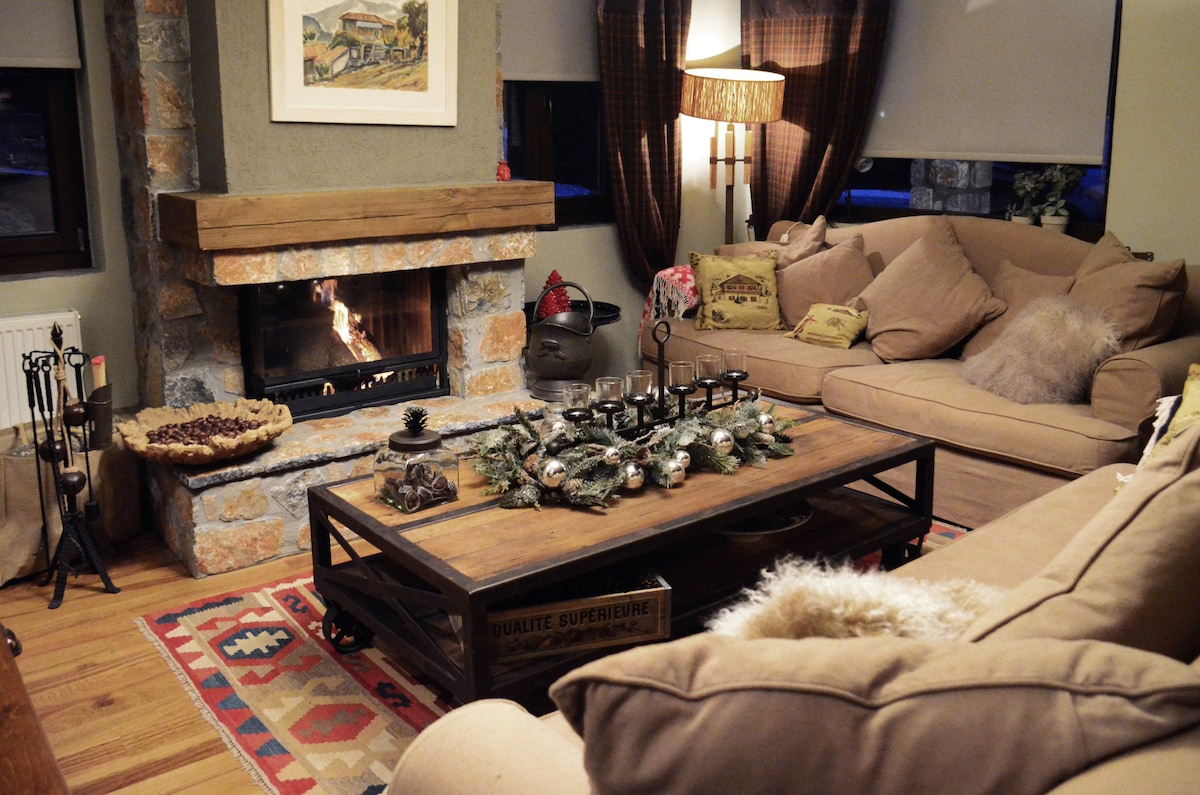A cozy living room is highlighted by a stone fireplace, with a soft sofa and armchairs arranged around a wooden coffee table. Decorative items and a plant are placed on the table, while warm lighting creates a welcoming ambiance.