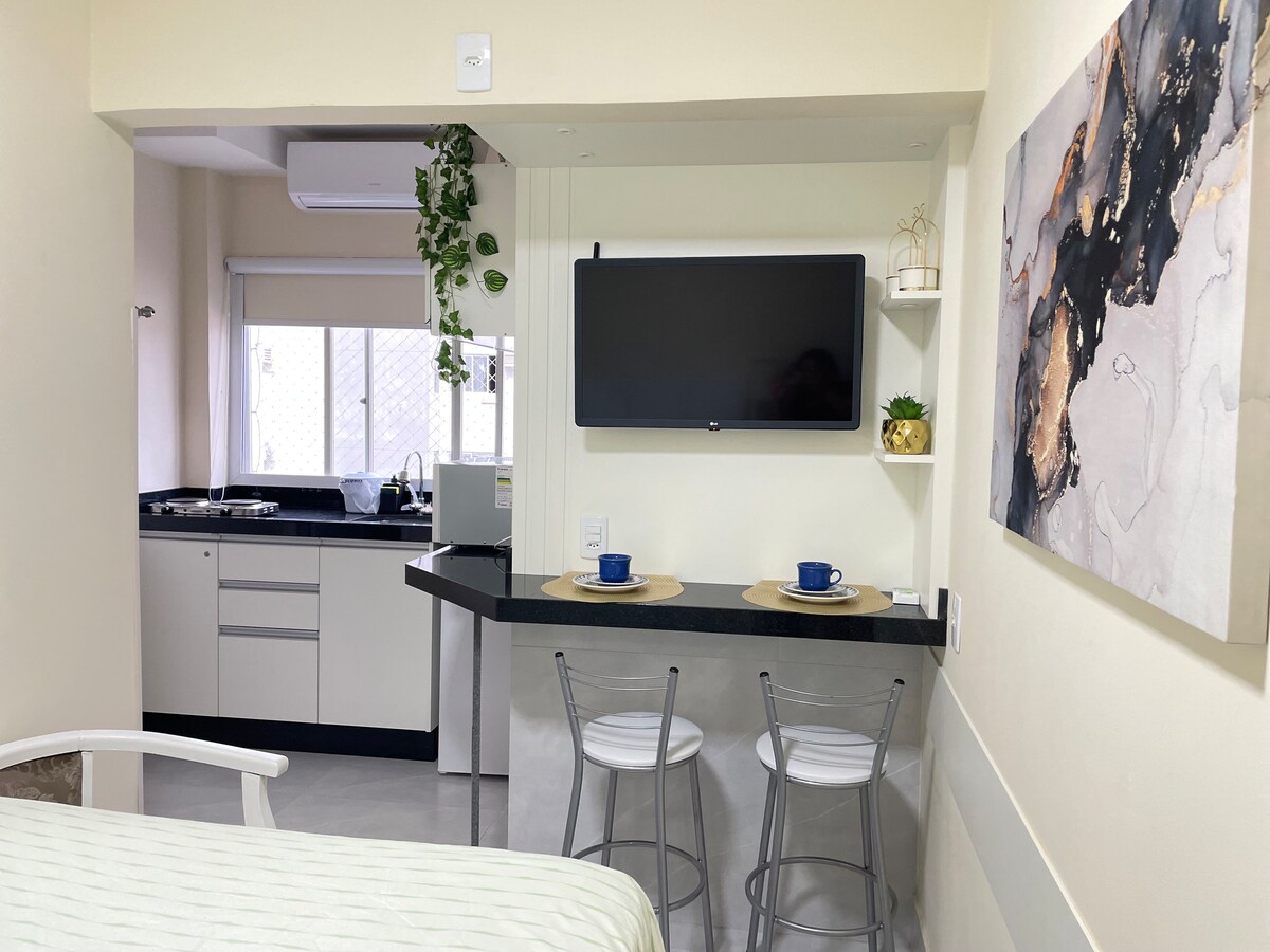 A compact living space displays a fully equipped kitchen in the background, featuring modern cabinetry and appliances. A cozy dining area with two clear chairs and blue cups is positioned in front of a wall-mounted TV. Soft natural light filters through the adjacent window.