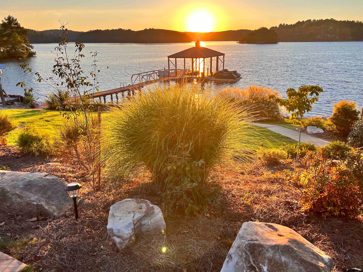 A serene lake view is captured at sunset, featuring a dock and pavilion extending into the water. Lush grasses and landscaped stones frame the scene, as warm sunlight bathes the area in golden tones, enhancing the tranquil atmosphere of the shoreline.