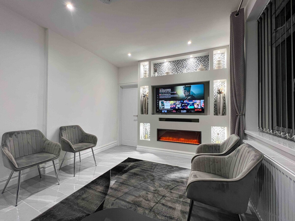 The living room features a media wall with a Smart 4K TV mounted above an electric fireplace. Two stylish, plush chairs are arranged on a contemporary rug, while soft lighting highlights the space and adds warmth to the neutral-toned walls.