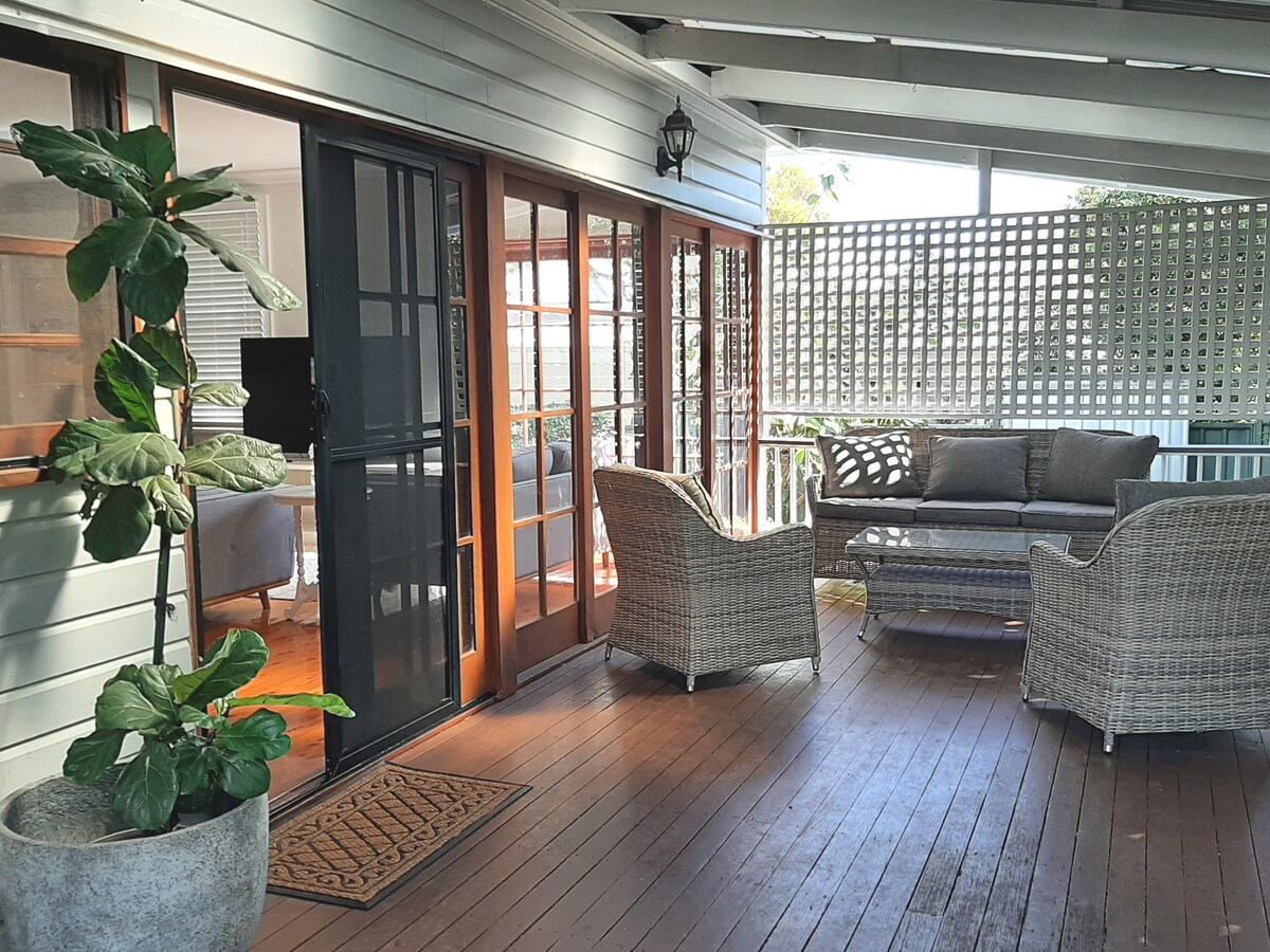 A covered porch area features a wooden deck with woven seating arranged around a low table. Double doors lead into the living space, while a decorative plant adds a touch of greenery. Latticework provides partial privacy and enhances the outdoor ambiance.