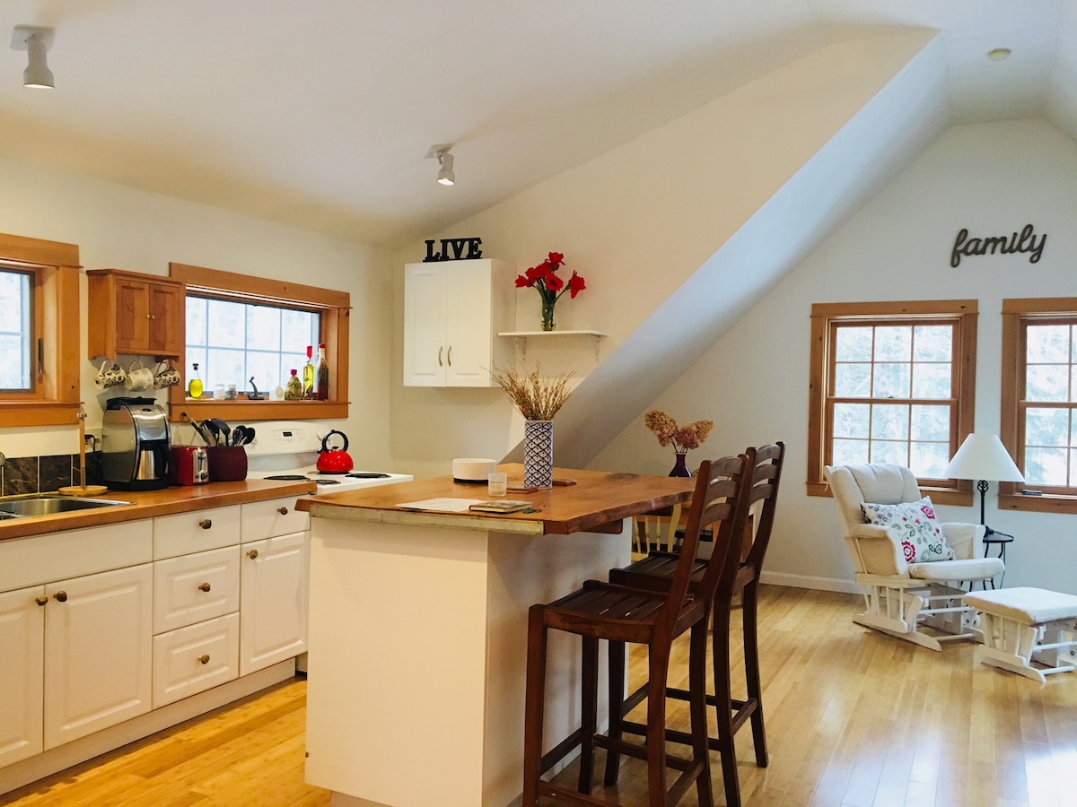 The kitchen area features white cabinetry and a wooden countertop, with modern appliances visible. Natural light enters through multiple windows, creating a bright space. A dining table with three chairs is positioned nearby, along with a cozy rocking chair and decorative elements on the walls.