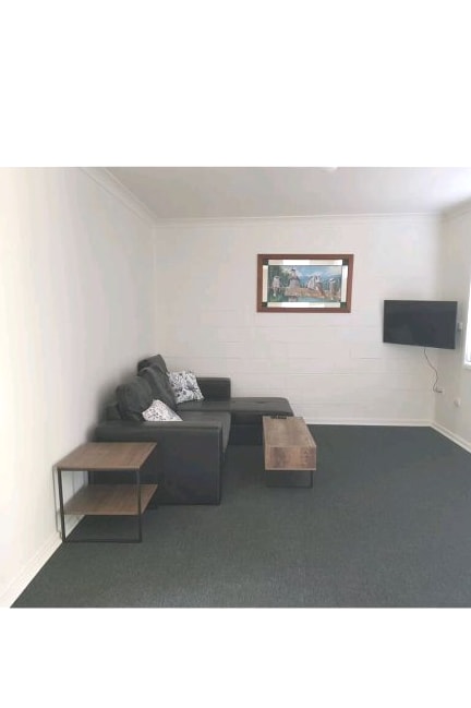 A comfortable living area features a gray sectional sofa adorned with patterned throw pillows. A wooden coffee table is positioned in front of the sofa, while a flat-screen television is mounted on the wall opposite. Natural light enters through a nearby window.