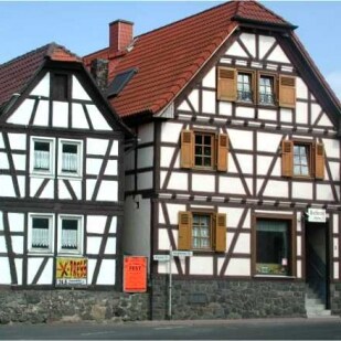 Two traditional half-timbered houses stand side by side, featuring wooden frameworks and decorative shutters. The building on the left displays a small sign, while both structures showcase warm-colored facades beneath a tiled roof. A clear sky provides a bright backdrop.