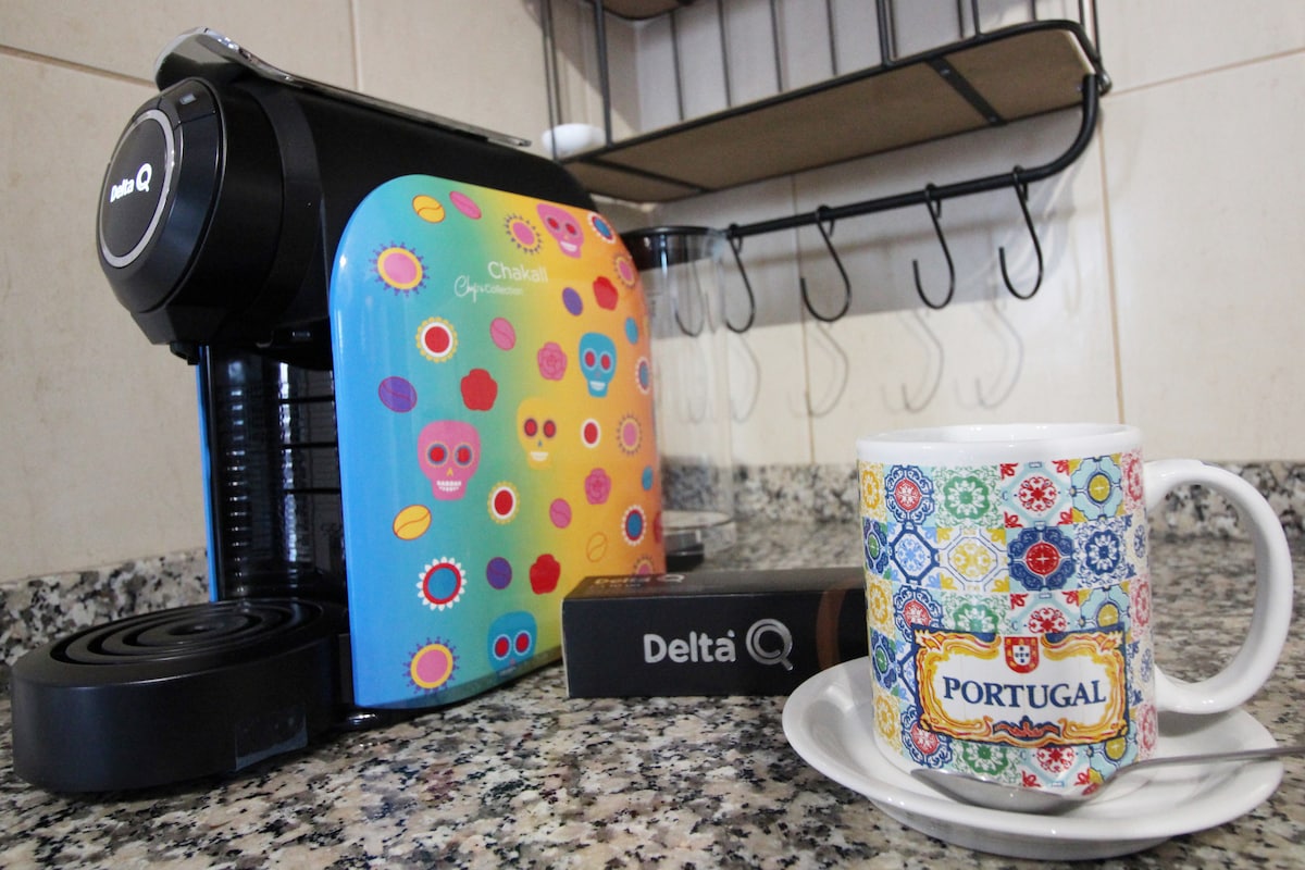 A colorful coffee machine with a bright, patterned cover is positioned on a speckled countertop. A decorative mug labeled 'PORTUGAL' rests on a saucer nearby, along with a box of coffee pods, offering a welcoming coffee experience.