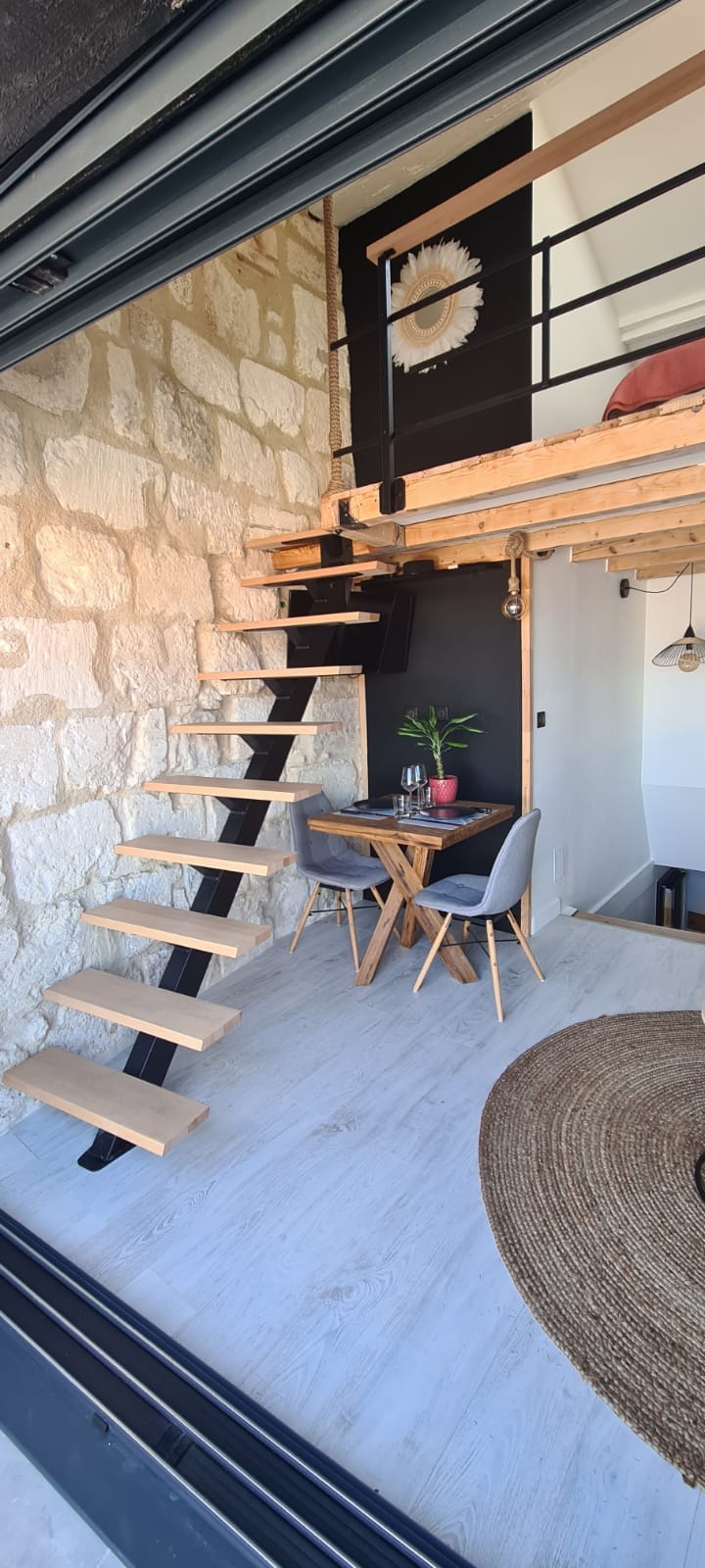 A wooden staircase leads to a mezzanine area, complemented by a stone wall. A small dining table with two chairs is positioned nearby, and a potted plant adds a touch of greenery. Natural light fills the space, enhancing the modern aesthetic.