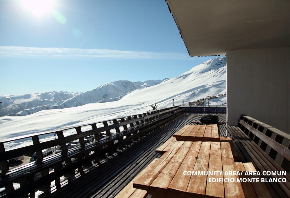 A communal area features a spacious wooden deck with long tables, providing open views of snow-covered mountains. Sunlight brightens the space, highlighting the clear skies above, ideal for relaxation and enjoying the surrounding natural beauty.