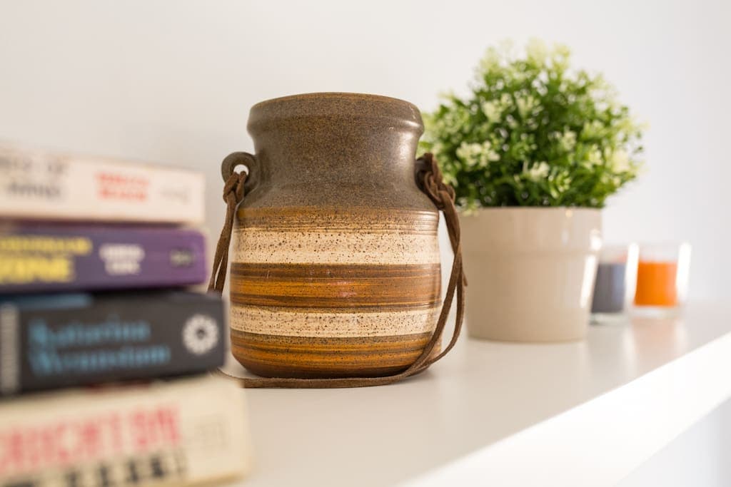 A decorative shelf is displayed, featuring a rustic brown and orange ceramic pot with a leather handle. Nearby, a cluster of books in various colors leans against the shelf, alongside a small potted plant. Soft candles in glass holders are positioned behind.
