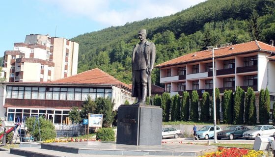 A bronze statue of a historical figure is prominently displayed in a plaza, surrounded by well-kept gardens and flowers. Multi-story buildings with terracotta roofs are positioned in the background, alongside a line of parked cars. Lush green hills are visible in the distance.