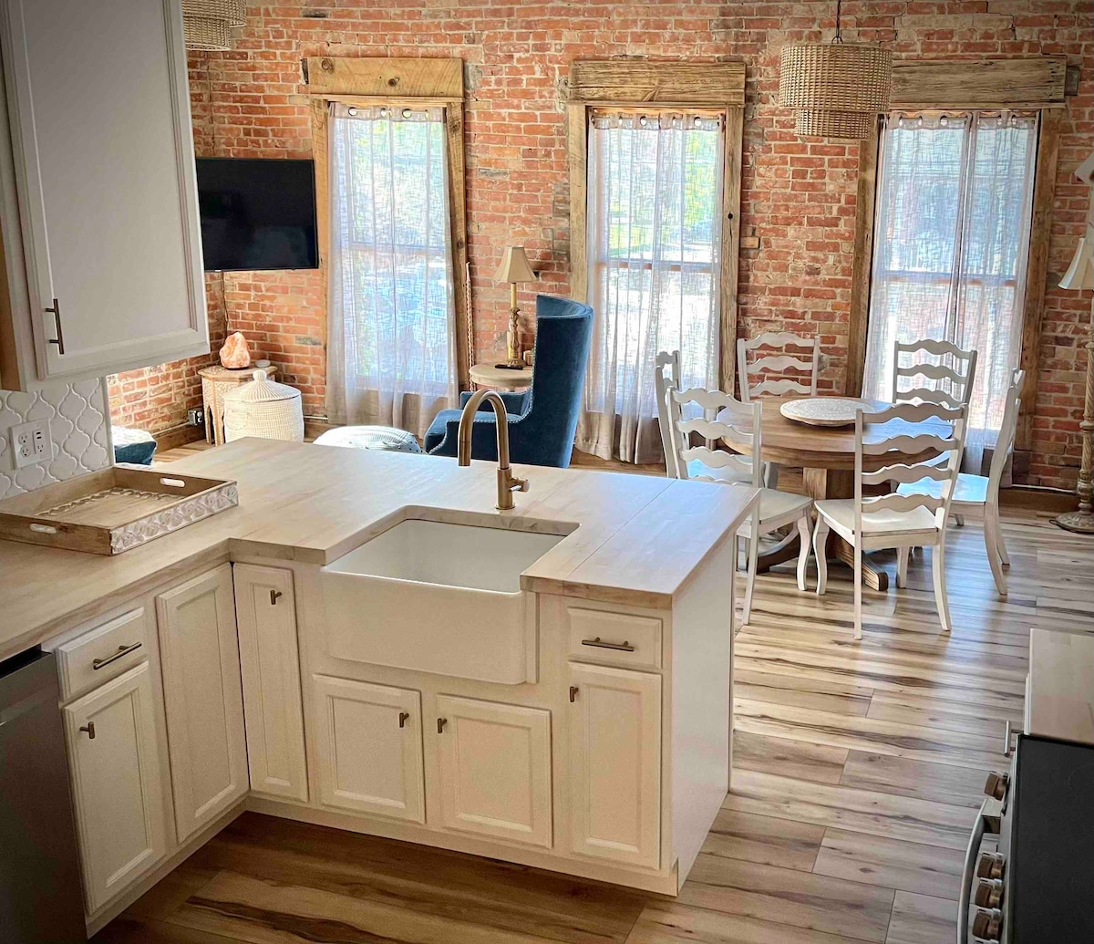 The kitchen area features a large farmhouse sink and a spacious island, surrounded by modern cabinetry. Exposed brick walls complement the light wood flooring. A cozy dining area is visible, showcasing a round table with white chairs and a comfortable blue armchair nearby. Large windows allow natural light to fill the space.