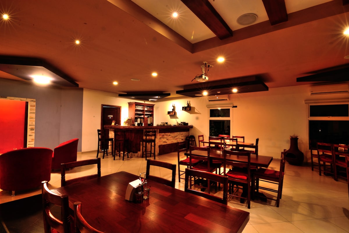 The interior of the restaurant features wooden tables and chairs arranged in a spacious layout. A bar area is visible in the background, with shelves displaying various beverages. Soft lighting enhances the warm atmosphere, while large windows provide glimpses of the outside.
