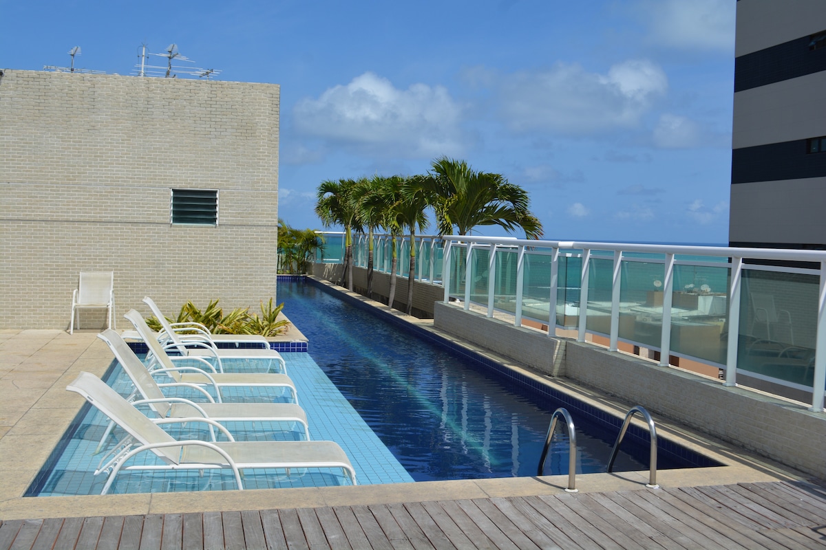 A rooftop swimming pool is surrounded by a sun terrace lined with white lounge chairs. Lush palm trees provide a tropical ambiance. The pool area offers unobstructed views of the tranquil sea, under a clear blue sky with a few scattered clouds.