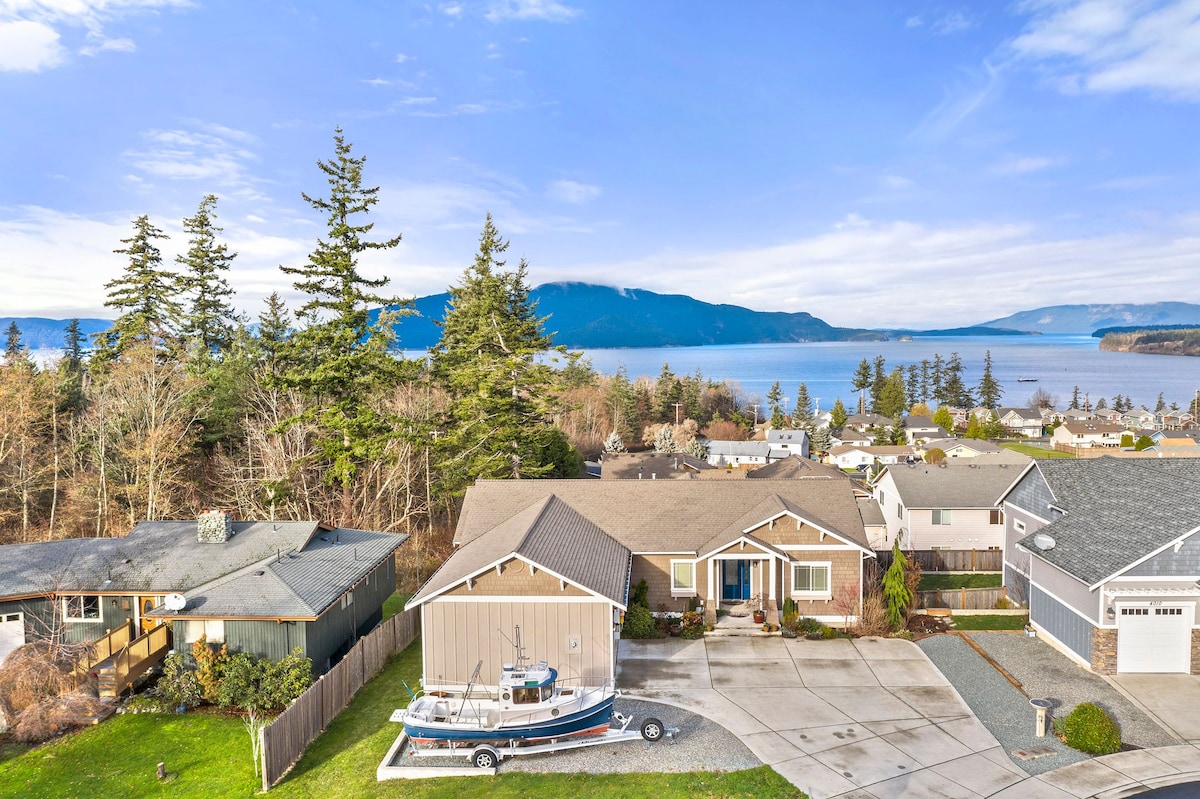 Aerial view of a Northwest-style home surrounded by natural scenery, including trees and a view of the water. A driveway and boat are visible in front of the house, with mountains rising in the background under a clear blue sky.