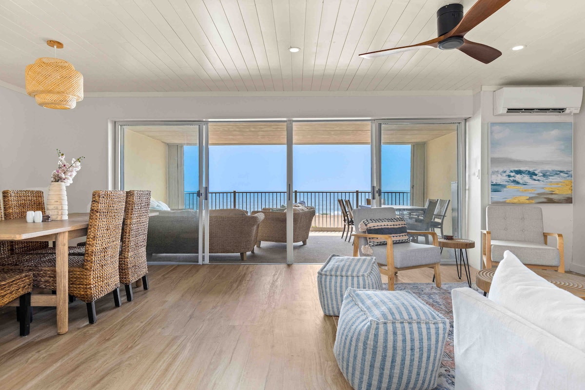 The open-plan living area features a dining table with woven chairs, a cozy seating arrangement, and large glass sliders that provide stunning ocean views. Light hardwood flooring complements the neutral decor, and artwork adorns one wall, enriching the coastal atmosphere.